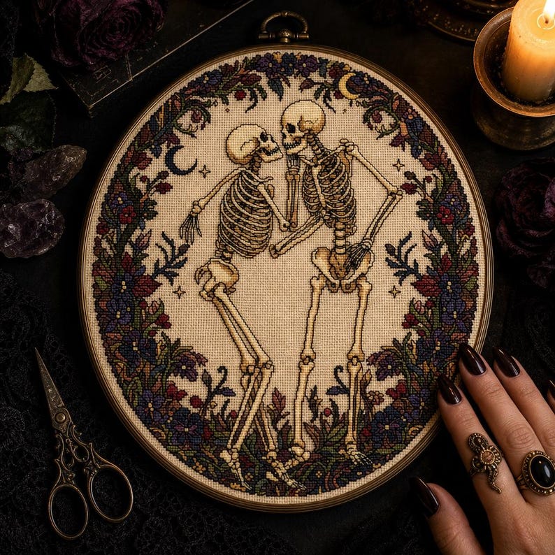 May include: A circular cross-stitch embroidery piece featuring two skeletons dancing, framed in a wooden hoop. The skeletons are surrounded by a detailed border of dark purple, red, and green flowers, leaves, and small crescent moon shapes on a beige fabric.