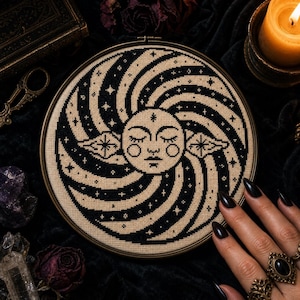 May include: A circular embroidery hoop containing a cross-stitch design of a central sun face with closed eyes, surrounded by black and beige swirling rays and small star motifs. The fabric is beige with black thread forming the detailed pattern.