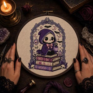 May include: A circular embroidery hoop containing a cross-stitch design of a small, hooded skeleton figure sitting on a stack of three books. The figure wears a purple cloak with a pink bow and holds a lantern. The scene is framed by purple vines.