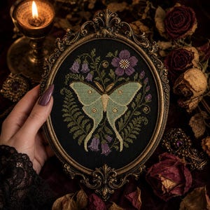 May include: A cross-stitch embroidery featuring a pale green moth centered on a black background, surrounded by purple flowers and green ferns. The artwork is encased in an ornate, oval-shaped, antique bronze-colored frame with intricate detailing.