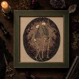 May include: A framed cross-stitch embroidery featuring two skeletons facing each other within an oval border of dark flowers and foliage. The skeletons are depicted in a light beige tone against a tan fabric background, surrounded by a dark green frame.