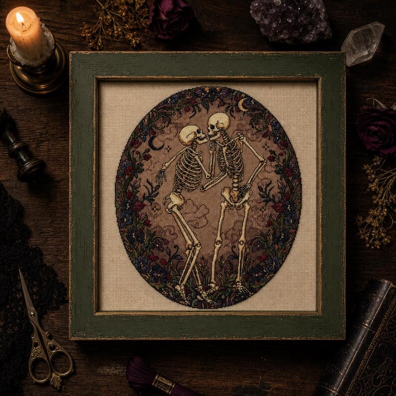 May include: A framed cross-stitch embroidery featuring two skeletons facing each other within an oval border of dark flowers and foliage. The skeletons are depicted in a light beige tone against a tan fabric background, surrounded by a dark green frame.