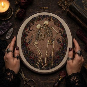 May include: A circular embroidery hoop containing a cross-stitch design of two skeletons dancing within a floral wreath. The skeletons are off-white, set against a tan fabric background with subtle cloud motifs, surrounded by dark red and blue flowers.