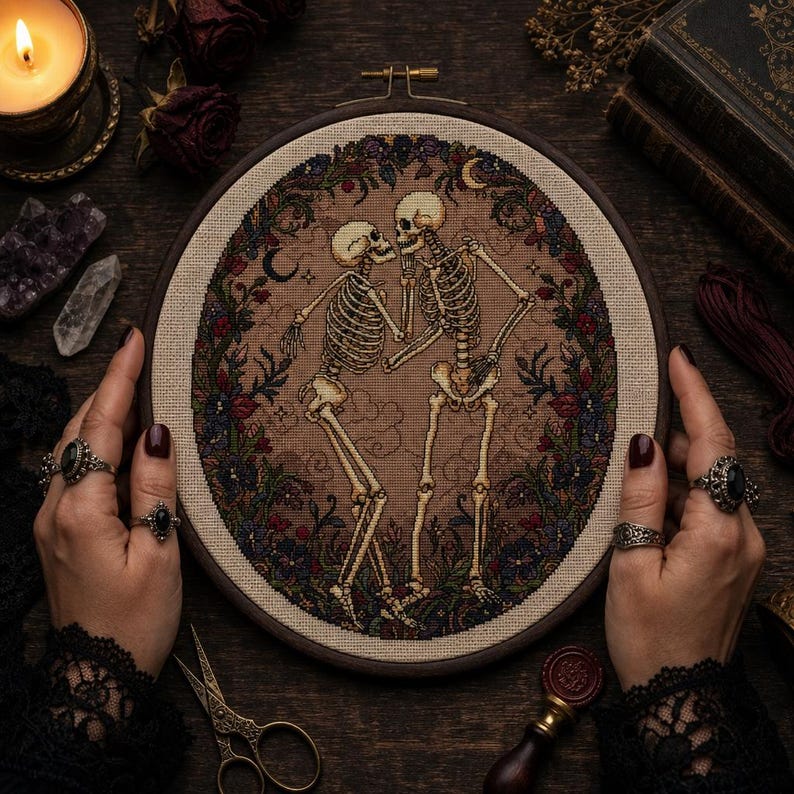 May include: A circular embroidery hoop containing a cross-stitch design of two skeletons dancing within a floral wreath. The skeletons are off-white, set against a tan fabric background with subtle cloud motifs, surrounded by dark red and blue flowers.