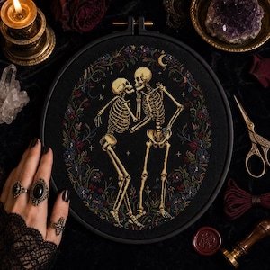 May include: A circular embroidery hoop containing a cross-stitch design on black fabric. The design features two beige skeletons facing each other, surrounded by a colorful floral wreath with small stars and a crescent moon in the background.