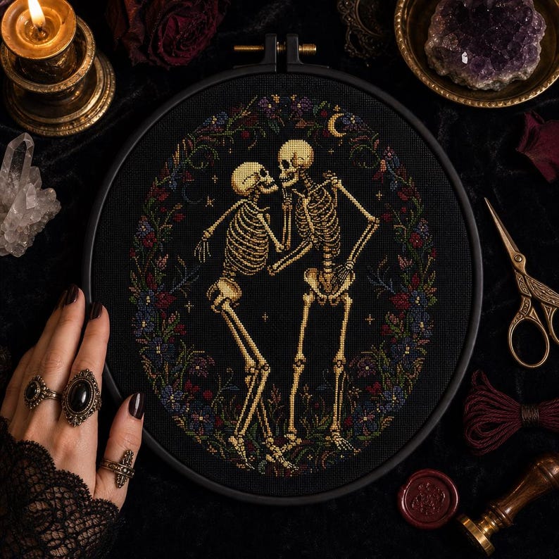 May include: A circular embroidery hoop containing a cross-stitch design on black fabric. The design features two beige skeletons facing each other, surrounded by a colorful floral wreath with small stars and a crescent moon in the background.