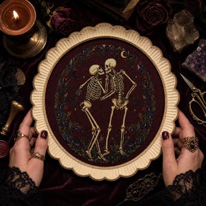May include: A circular cross-stitch embroidery featuring two skeletons dancing within a dark maroon floral wreath. The piece is encased in a scalloped, cream-colored frame. The design includes small crescent moons and stars against a deep red fabric.