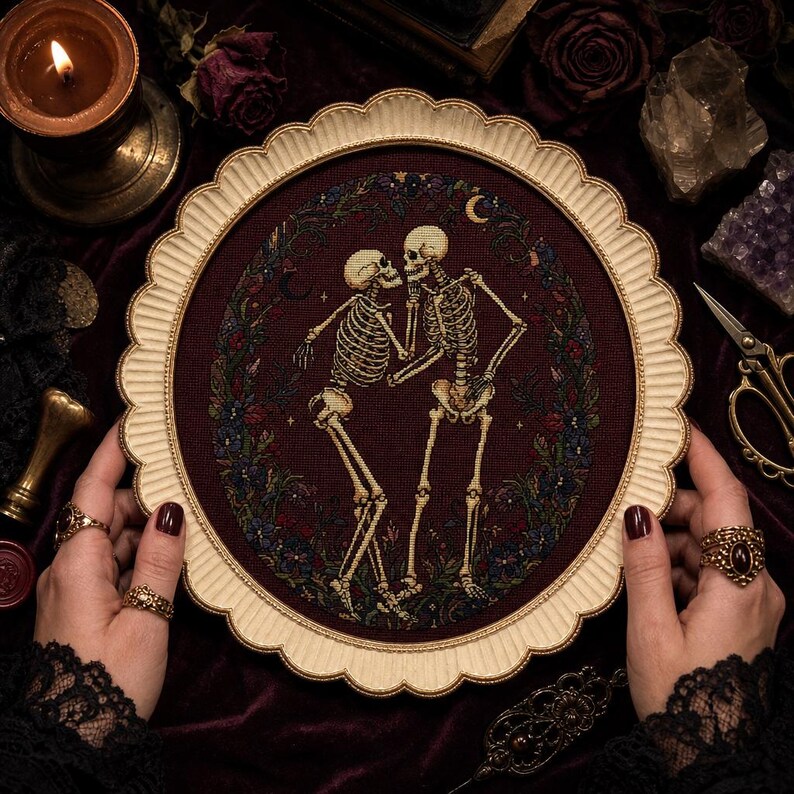 May include: A circular cross-stitch embroidery featuring two skeletons dancing within a dark maroon floral wreath. The piece is encased in a scalloped, cream-colored frame. The design includes small crescent moons and stars against a deep red fabric.
