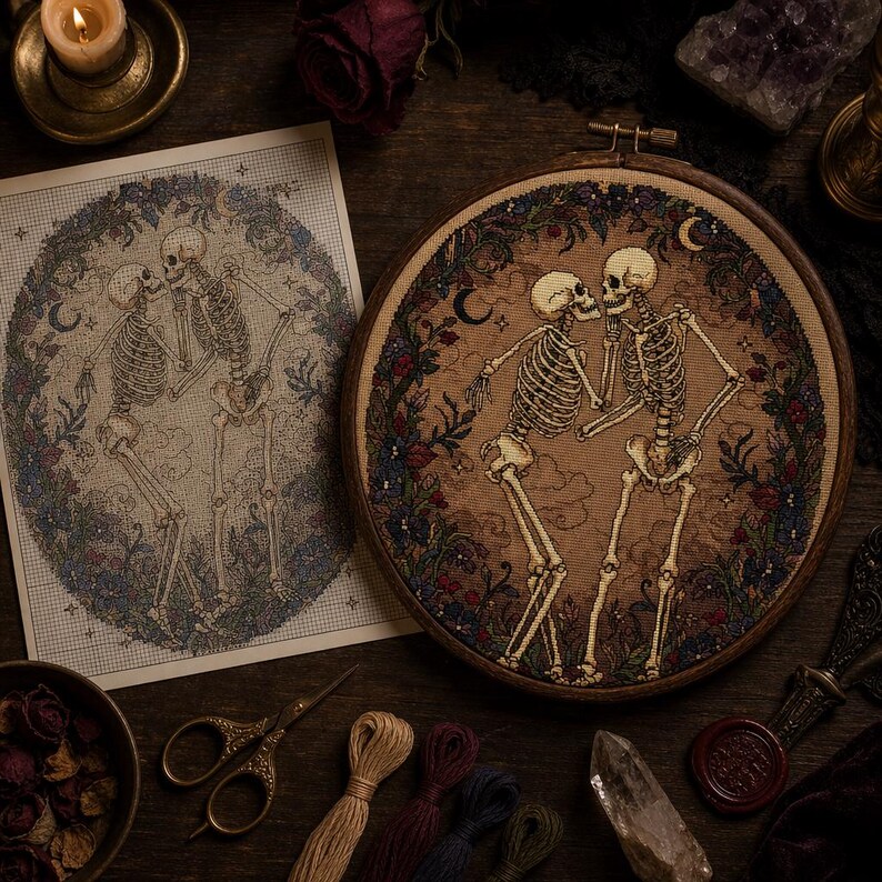 May include: A circular wooden embroidery hoop containing a cross-stitch design of two skeletons facing each other, surrounded by a floral wreath with crescent moons. Beside it lies a printed paper pattern of the same skeleton design.