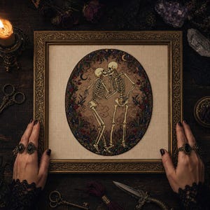 May include: A framed cross-stitch embroidery featuring two skeletons dancing within an oval border of dark flowers and crescent moons. The skeletons are depicted in a beige-toned fabric setting, enclosed in an ornate, antique gold-colored frame.