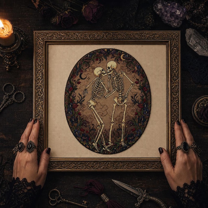 May include: A framed cross-stitch embroidery featuring two skeletons dancing within an oval border of dark flowers and crescent moons. The skeletons are depicted in a beige-toned fabric setting, enclosed in an ornate, antique gold-colored frame.