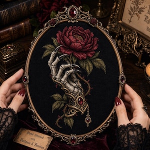 May include: An oval-framed cross-stitch embroidery on black fabric featuring a skeletal hand with long, dark-tipped fingers holding a large, deep red rose with green leaves. The ornate, metallic gold frame is embellished with small, dark red gemstones.