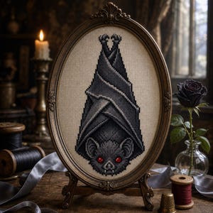 May include: An oval-framed cross-stitch embroidery featuring a gray bat hanging upside down with bright red eyes, displayed in an ornate bronze-colored frame. The needlework is stitched on light beige fabric, showcasing detailed shading and texture.