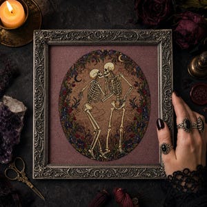 May include: A framed cross-stitch embroidery featuring two skeletons standing face-to-face within an oval floral wreath. The skeletons are depicted in a beige tone against a dark mauve fabric background, surrounded by intricate dark metal frame.
