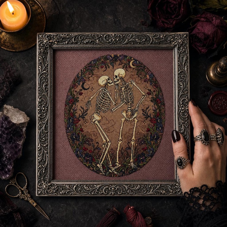 May include: A framed cross-stitch embroidery featuring two skeletons standing face-to-face within an oval floral wreath. The skeletons are depicted in a beige tone against a dark mauve fabric background, surrounded by intricate dark metal frame.