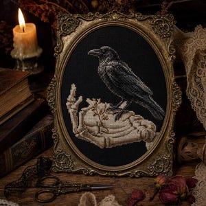 Könnte beinhalten: Eine Kreuzstichstickerei mit einem schwarzen Raben, der auf einer skelettierten Hand sitzt, in einem verzierten, ovalen antiken Goldrahmen. Das Design verwendet schwarzes, weißes und beiges Garn, um den Vogel und die Knochen auf schwarzem Stoff darzustellen.