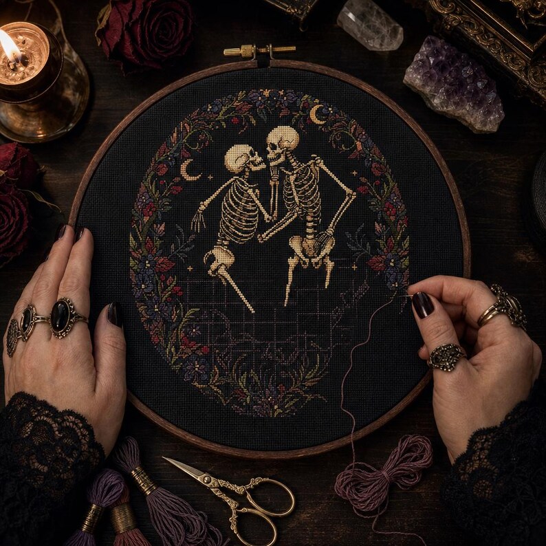 May include: A circular wooden embroidery hoop containing a cross-stitch design on black fabric. The design features two beige skeletons facing each other, surrounded by a detailed floral wreath in shades of red, purple, and green with small crescent moons.
