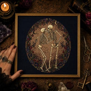 May include: A framed cross-stitch embroidery featuring two skeletons facing each other in a dance pose. The skeletons are set against a tan oval background surrounded by a dark blue border with floral motifs, small stars, and crescent moons.