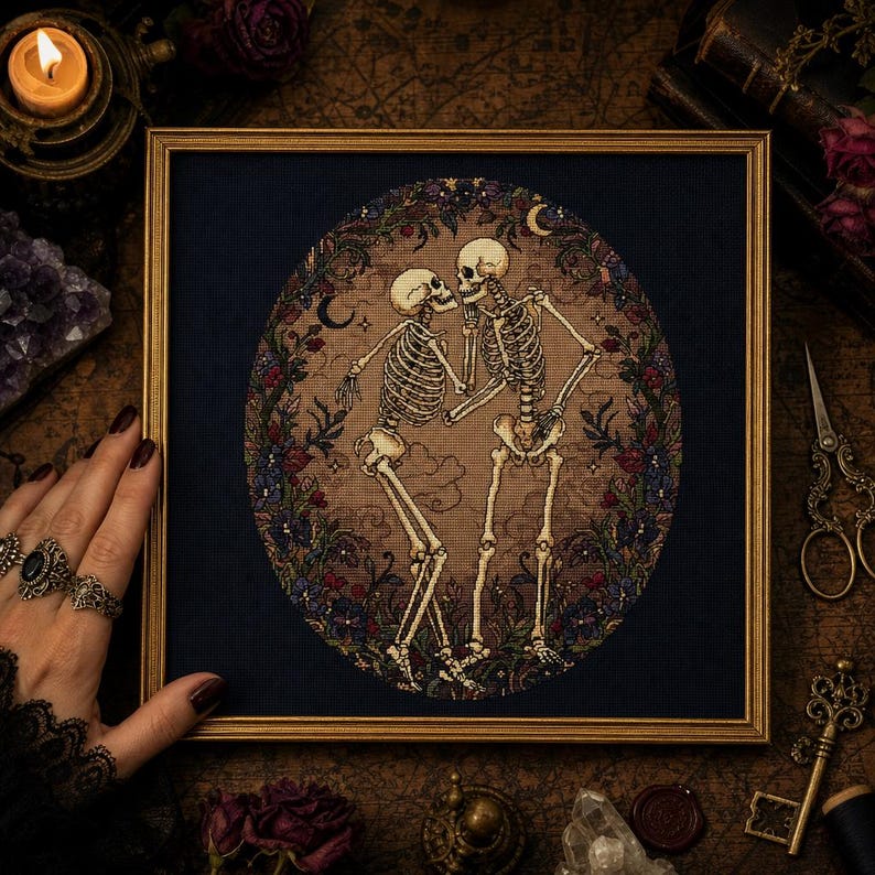 May include: A framed cross-stitch embroidery featuring two skeletons facing each other in a dance pose. The skeletons are set against a tan oval background surrounded by a dark blue border with floral motifs, small stars, and crescent moons.