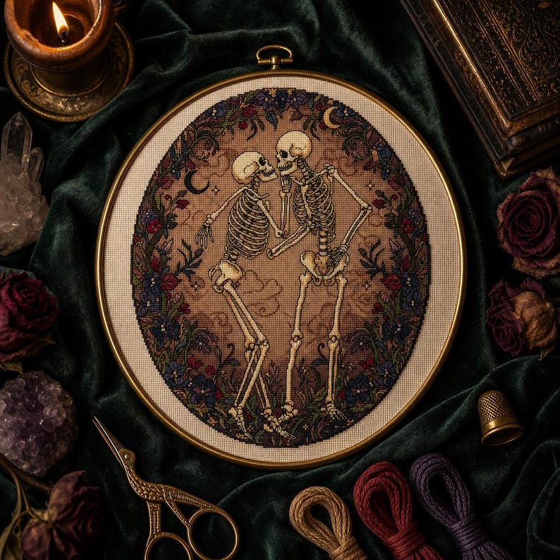 May include: A circular embroidery hoop containing a cross-stitch design of two skeletons facing each other, surrounded by a dark floral wreath with crescent moons. The skeletons are light beige, set against a tan background with cloud motifs.