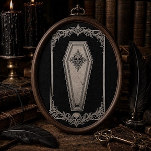 May include: An oval-framed cross-stitch embroidery featuring a light-colored coffin centered on a black fabric background. The coffin is adorned with a skull emblem, and the entire piece is surrounded by an intricate, decorative border with a skull.