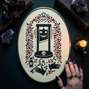 May include: An oval cross-stitch embroidery in a gold hoop featuring a black guillotine with a smiling face, surrounded by a floral border of red roses and black vines. Below the guillotine are a skull, a potion bottle, and two black hands.