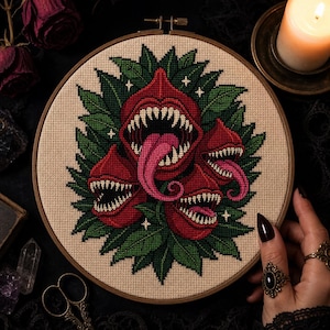 May include: A circular wooden embroidery hoop containing a cross-stitch design of three red, toothy, carnivorous plants with pink tongues and green leaves on a beige fabric background, accented with small white four-pointed star shapes.
