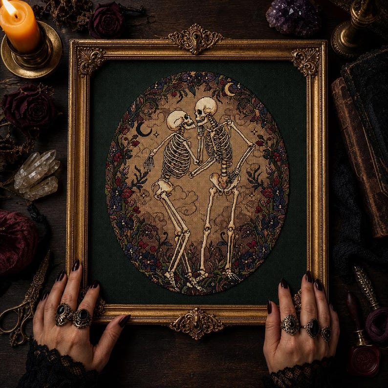 May include: A framed cross-stitch embroidery featuring two skeletons dancing within an oval border of dark flowers and crescent moons. The artwork is set against a dark green fabric background and enclosed in an ornate, vintage-style gold frame.