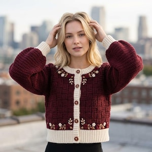 Könnte beinhalten: Eine tiefburgunderrote Strickjacke mit Korbgeflecht-Textur, cremefarbenen gerippten Bündchen, Saum und Knopfleiste. Das Kleidungsstück ist mit cremefarbenen und burgunderroten Blumenstickereien verziert und wird mit fünf kleinen, runden, dunklen Knöpfen geschlossen.