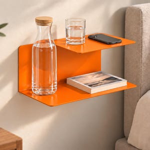 Könnte beinhalten: Ein leuchtend orangefarbenes Wandregal aus Metall mit zwei Ebenen. Auf der oberen Ebene stehen eine durchsichtige Glaswasserflasche mit Holzverschluss und ein Glas Wasser, auf der unteren Ebene liegt ein rechteckiges Buch mit Landschaftsbild.