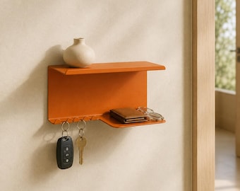 May include: A wall-mounted orange metal shelf featuring a flat top surface, a lower section with integrated hooks for hanging items, and a small side platform. A small beige vase, a brown wallet, and metal-rimmed glasses rest on the shelf.