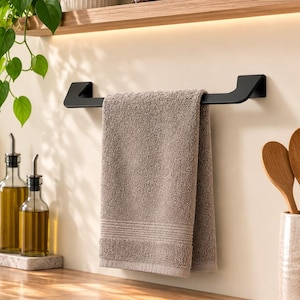 Puede incluir: Un toallero de metal negro mate montado en la pared, que sostiene una toalla de cocina de algodón de color marrón claro doblada. El toallero presenta un diseño angular y elegante, situado sobre una encimera de madera en una cocina.