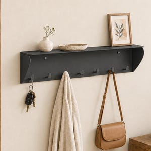 Peut inclure: Une étagère murale en métal noir mat dotée d'une surface plane et d'une rangée de six crochets intégrés. L'étagère supporte un petit vase blanc, un plat peu profond et une impression botanique encadrée. Des clés, une serviette beige et un sac beige y sont suspendus.