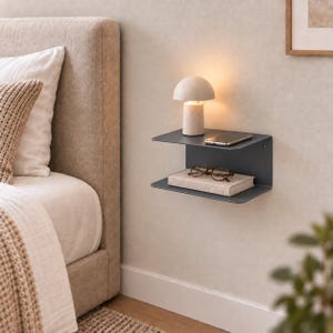 Op de afbeelding: Een donkergrijze metalen wandplank met twee niveaus. Het bovenste niveau bevat een kleine paddenstoelvormige lamp en een smartphone. Het onderste niveau bevat een gesloten boek met een bril met schildpadmontuur bovenop de kaft.