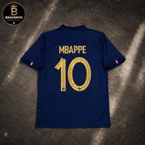 Puede incluir: Una camiseta de manga corta azul oscuro con el nombre MBAPPE y el número 10 impresos en dorado en la parte posterior. Las mangas tienen pequeños detalles con los colores azul, blanco y rojo. Un logo circular con el texto BRAVENTO es visible.