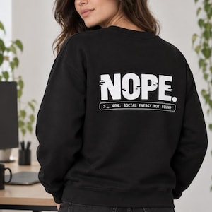 Op de afbeelding: Een zwart sweatshirt met lange mouwen en een ronde hals, voorzien van witte tekst op de achterkant die NOPE. leest, gevolgd door een rechthoekig kader met de tekst >_ 404: SOCIAL ENERGY NOT FOUND. Het sweatshirt heeft geribbelde boorden.