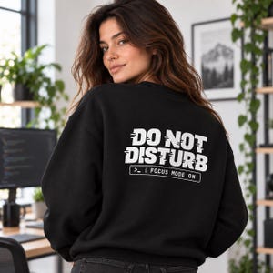 Puede incluir: Una sudadera negra de manga larga con texto blanco en la espalda que dice DO NOT DISTURB con un efecto de glitch, seguido de un recuadro rectangular que contiene el texto >_ | FOCUS MODE ON en una fuente sans-serif.