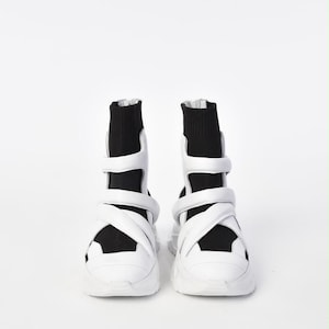 May include: A pair of high-top sneakers featuring a black sock-like upper and a white rubber sole. The exterior is accented with multiple white straps that wrap around the midfoot and ankle, secured with a vertical zipper at the back of the heel.