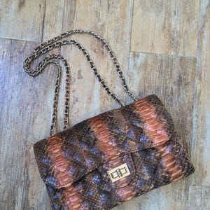 Puede incluir: Un bolso de hombro rectangular con un patrón de piel de serpiente texturizado en tonos marrón, bronceado y gris oscuro. El bolso incluye un cierre de giro de metal dorado en la solapa frontal y una correa de hombro de cadena de metal a juego.