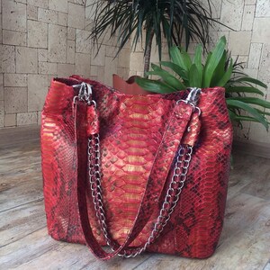 May include: A tote bag featuring a vibrant red and gold snakeskin pattern with a textured finish. The bag includes two shoulder straps, each constructed with a combination of matching red material and polished silver-tone metal chain links.