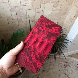 Peut inclure: Un portefeuille rectangulaire pr&eacute;sentant un motif de peau de serpent rouge vif et cramoisi fonc&eacute; avec une surface textur&eacute;e et &eacute;cailleuse. Le portefeuille comprend une fermeture &eacute;clair en m&eacute;tal dor&eacute; sur le c&ocirc;t&eacute; pour le compartiment principal.