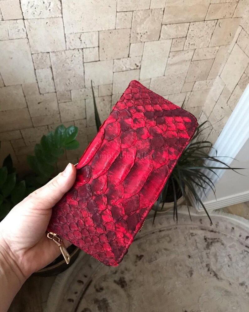 Peut inclure: Un portefeuille rectangulaire pr&eacute;sentant un motif de peau de serpent rouge vif et cramoisi fonc&eacute; avec une surface textur&eacute;e et &eacute;cailleuse. Le portefeuille comprend une fermeture &eacute;clair en m&eacute;tal dor&eacute; sur le c&ocirc;t&eacute; pour le compartiment principal.