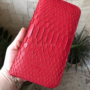 May include: A vibrant red rectangular wallet featuring a textured snakeskin pattern across the entire exterior surface. The wallet is secured with a silver-toned metal zipper pull located at the bottom corner, providing a functional closure mechanism.