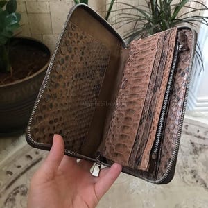 May include: A brown snakeskin textured wallet with a full wrap-around silver metal zipper. The interior features multiple card slots, a central zippered compartment, and a large open pocket, all lined with the same patterned snakeskin material.