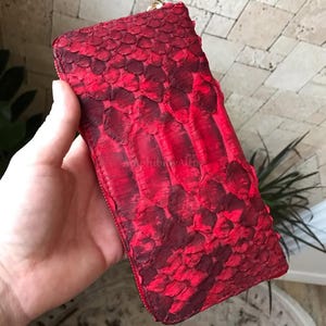Peut inclure: Un portefeuille rectangulaire pr&eacute;sentant une texture en peau de serpent rouge vif et bordeaux fonc&eacute; avec des &eacute;cailles superpos&eacute;es. Le portefeuille est s&eacute;curis&eacute; par une fermeture &eacute;clair en m&eacute;tal dor&eacute; le long du bord sup&eacute;rieur.