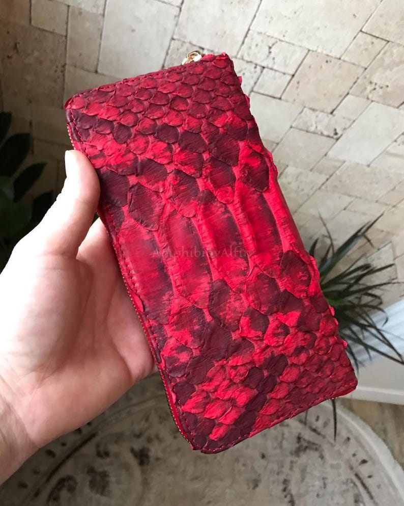 Peut inclure: Un portefeuille rectangulaire pr&eacute;sentant une texture en peau de serpent rouge vif et bordeaux fonc&eacute; avec des &eacute;cailles superpos&eacute;es. Le portefeuille est s&eacute;curis&eacute; par une fermeture &eacute;clair en m&eacute;tal dor&eacute; le long du bord sup&eacute;rieur.