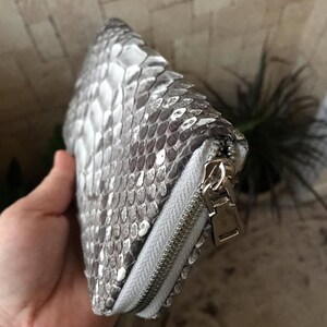 Op de afbeelding: Een rechthoekige portemonnee met een gestructureerd slangenleerpatroon in zilver en grijs. De portemonnee is voorzien van een zilverkleurige metalen ritssluiting langs de zijkant, inclusief een bijpassende zilverkleurige metalen ritstrekker.