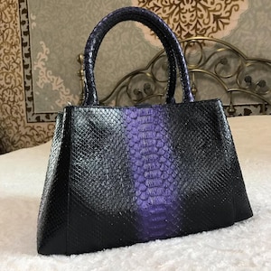 Könnte beinhalten: Eine strukturierte Handtasche mit strukturiertem Schlangenledermuster, tiefschwarzen Seiten und einem leuchtend violetten vertikalen Streifen in der Mitte. Die Tasche verfügt über zwei passende, abgerundete Tragegriffe an der Oberseite.