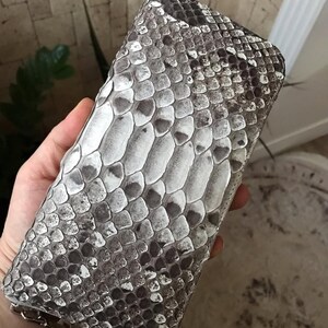 Op de afbeelding: Een rechthoekige portemonnee met een gestructureerd slangenleerpatroon in tinten wit, grijs en donkerbruin. Het oppervlak vertoont overlappende, schubachtige details over de gehele buitenkant, met een zilverkleurige metalen ritssluiting aan de onderkant.