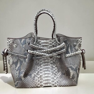 May include: A handbag featuring a grey and white snakeskin pattern with a textured scale finish. The bag includes two top handles, a central knotted strap detail across the front, and side ties with knotted ends, all crafted in a matching snakeskin material.
